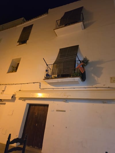This image shows the exterior of a building at night in Ibiza, Spain. The building is white and appears to be several stories high. There are multiple windows and balconies visible, some with wooden shutters. One balcony has a plant and an orange pillow on it. There is a wooden door visible at ground level, and a dark-colored chair is partially visible in the foreground. Above the door, there is a sign with cursive writing that appears to say "Central Ibiza". Wires and conduit are running along the exterior wall. The lighting is warm and artificial, suggesting artificial lighting is illuminating the scene.