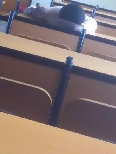 The image shows a person sleeping face down on a desk in a classroom. The person is wearing a white shirt and has dark, curly hair. The desk is made of wood and has black metal legs. The classroom appears to be empty except for the sleeping person. The lighting suggests it could be daytime. There is no visible text in the image. The overall impression is one of fatigue or disinterest in a learning environment.