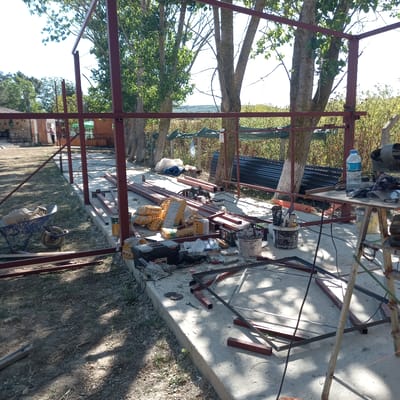 The image depicts an outdoor construction site in Çatalca, Turkey, characterized by a partially constructed metal frame structure. Red-painted steel beams form the primary structural elements of a shed or similar building. The ground surface is a mix of concrete and soil, with scattered construction materials.

Foreground elements include a blue wheelbarrow containing bags of construction material, various metal beams, a black toolbox, and a white bucket filled with tools. Bags labeled "FIXA" are prominently stacked, suggesting they contain cement or similar building supplies. A partially assembled metal frame lies on the concrete, alongside a wrench and cutting discs.

The middle ground shows more steel beams arranged on the concrete foundation, and a collection of buckets and tools. A water bottle sits on a makeshift table, next to what appears to be a welding machine. In the background, a line of trees with green foliage creates a natural boundary, with the bright sky visible through the branches. Further back, a building with wooden cladding and another structure with a green awning are partially visible. A person in a light-colored shirt and dark pants can be seen standing near the entrance of the wooden building. The overall scene suggests an active but paused construction project under daylight.