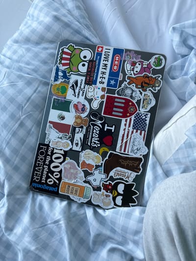 The image shows a laptop covered in stickers on top of a white and blue gingham fabric. The laptop is cluttered with a variety of stickers. Some notable stickers include "H-E-B", "Nashville", "I Heart Mozart's Coffee Roasters Lake Austin, Texas", "Dazed and Confused", "Nothing But Rock N Roll", an American flag, a Mexican flag, and "100% for the planet forever". Mozart's Coffee Roasters is a popular coffee shop located on Lake Austin in Austin, Texas.