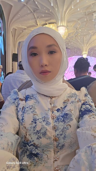 In Karakol City, Kyrgyzstan, a young woman wearing a white hijab and a floral-patterned jacket with blue and white blossoms is seated indoors. She is looking directly at the camera. The background features an ornate ceiling with illuminated crystal-like decorations and architectural arches. Other individuals are visible in the background, some facing away from the camera. A large screen displaying an image is to the left, and to the right, a pink banner with text is partially visible. The overall environment appears to be an indoor event or venue.