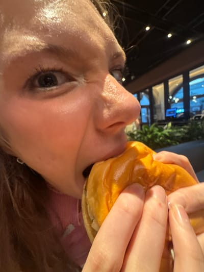 A close-up shot captures a young woman biting into a hamburger. Her face is partially obscured by the burger, with her eyes wide open and a slight grimace of enjoyment or effort. The hamburger bun is golden brown and appears glossy, held by her hands. The background is blurred, showing an indoor setting with a dark ceiling with lights and a view of windows with plants and lights visible outside, suggesting a restaurant or dining area. The image does not contain any information that would indicate its location in Pudong, China.