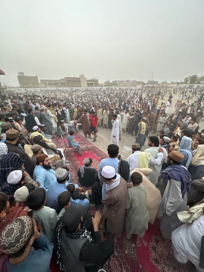 This media file depicts a large gathering of people, predominantly men and boys, engaged in a religious prayer or sermon. The scene appears to be outdoors, with a dusty or hazy atmosphere, possibly indicating a hot and dry climate. The ground is covered with rugs and a sandy or dirt surface. Many individuals are wearing traditional Islamic attire, including shalwar kameez and kufi caps. Some are seen kneeling or prostrating in prayer, while others are seated.
The gathering is extensive, stretching far into the background, suggesting a significant communal event. The architecture visible in the background indicates an urban or semi-urban setting, possibly within a residential or open area. The overall mood seems solemn and devout, as people participate in their religious observance. The time of day is difficult to ascertain precisely due to the atmospheric conditions, but the light suggests it could be morning or late afternoon. No visible text is present in the image. The location context of Chaman City Tehsil, Pakistan, further supports the interpretation of a religious gathering in a region with Islamic cultural practices.