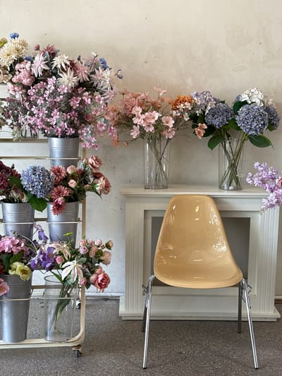 The image depicts a display of artificial flowers in a retail setting, possibly a flower shop or a home decor store, located in Tam Kỳ, Vietnam. The background features a textured, pale wall. In the foreground, a modern, light-tan molded plastic chair with chrome legs sits on a grey speckled floor.
To the left of the chair, a multi-tiered metal cart holds several buckets of artificial flowers. The top shelf has a large arrangement of white, pink, and blue flowers. The middle shelf displays buckets with various flower arrangements, including blue hydrangeas and pink dahlias. The bottom shelf contains more flower buckets, one with pink and purple flowers.
To the right of the chair, a white, decorative stand or mantlepiece is visible. On top of this stand, several glass vases hold different bouquets of artificial flowers. These include pink blossoms, orange flowers mixed with purple and blue hydrangeas, and white hydrangeas. To the far right, a sprig of purple artificial flowers extends into the frame. The overall impression is a curated display of floral arrangements for sale.