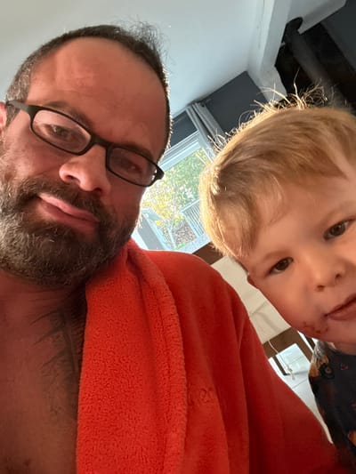 A selfie taken from a low angle features a man with glasses and a beard in the foreground and a young child with blond hair looking down towards the camera in the background. The man is wearing an orange garment and appears to be indoors. A window with trees visible outside can be seen in the background. The location, Marche-en-Famenne, Belgium, cannot be determined from the visual information provided.