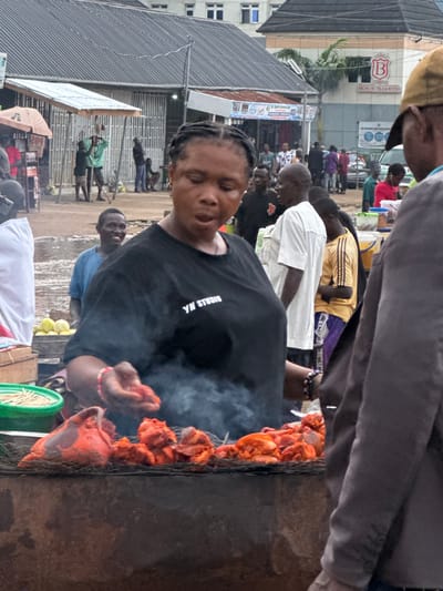 An outdoor market scene is depicted in Obinze, Nigeria, with a focus on a woman grilling meat. The woman, positioned in the foreground, wears a black t-shirt with white lettering that reads "YN STUDIO." She is actively tending to pieces of raw, red meat arranged on a grill, which is emitting smoke. Her right hand is visible, appearing to be in the process of handling the meat.
Surrounding the grilling station, other individuals are present. To the left of the woman, a person wearing a white shirt is partially visible. Behind the grilling station, a young man in a blue t-shirt is looking towards the camera and smiling. Further in the background, other people are seen walking and standing, some near market stalls. One stall has a sign with the "B" logo of "BRUNO HOTELS & SUITES."
The environment suggests a typical Nigerian street market with various structures, including corrugated metal roofs and awnings. The ground appears to be dirt or gravel. The overall atmosphere is busy and dynamic, characteristic of an active marketplace.