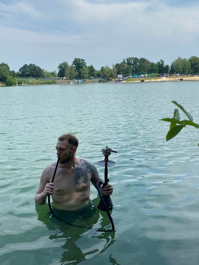 A shirtless man with tattoos on his left arm and chest is standing in shallow, greenish water, holding a hookah pipe. The water is rippling, and a portion of the hookah's stem and hose are visible. In the background, across the water, is a sandy beach area with trees, some buildings, and a clear sky with some clouds. The scene appears to be a recreational area, possibly a lake or large pond, within the city of Kaniów, Poland.