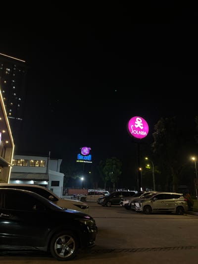 An exterior nighttime shot shows a parking lot in front of a commercial building in the city of South Tangerang, Indonesia. The building has a modern facade with large windows and an illuminated awning. To the left, a tall residential building is visible. Two large, illuminated signs are prominent in the scene: one is a pink circular sign with the word "SOLARIA" and a stylized icon, and the other is a neon blue sign with the words "ME" and "Gacoan". Several cars are parked in the lot, including a black SUV in the foreground. Streetlights illuminate the area, casting a warm glow on the pavement and trees. A red sign with Indonesian text is visible in the background, likely a warning or information sign. The sky is dark, indicating it is nighttime.