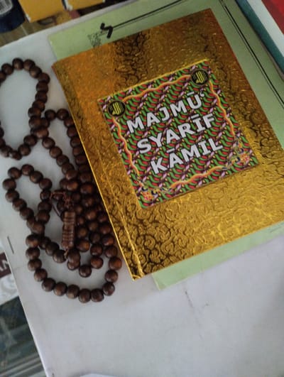 The image shows a close-up view of a gold-covered book with Arabic calligraphy on its cover, placed on a light green book. To the left of the gold book is a string of dark brown prayer beads. The background is blurred but appears to be a flat, white surface with some faint markings. The overall scene suggests a religious or spiritual context, possibly a personal collection of religious items. The image lacks any specific details to place it in Tanjungsari, Indonesia, or any other city.