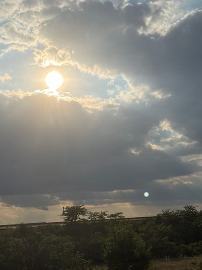 The media file shows a daytime scene in Saraia, Bulgaria, featuring a sky filled with dramatic clouds and the sun. The sun is prominently visible, appearing as a bright orb peeking through or behind the clouds, casting rays of light. There are two distinct areas of sunlight captured in the image. One is a large, bright sun with rays extending outwards, partially obscured by large, textured clouds. The second, smaller sun-like object is visible further up in the sky, appearing as a distinct, bright circle against a darker, cloudy backdrop.
The lower portion of the image reveals a landscape with dense green foliage, suggesting trees and other vegetation. A thin, dark line runs horizontally across the frame, potentially indicating a fence, wall, or road, separating the sky from the ground. The overall lighting suggests it is either early morning or late afternoon due to the quality of light and the position of the sun. The weather appears to be partly cloudy, with a mix of dark and bright cloud formations. There are no people or distinct man-made objects clearly visible other than the implied barrier at the bottom. The scene evokes a sense of natural beauty and perhaps tranquility, with the interplay of light and shadow in the sky.