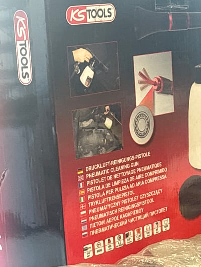 The image shows a close-up of a product box, likely for a tool. The box features prominent branding with "KS TOOLS" in red and white lettering. Various product-related imagery and text are visible, including what appears to be a pneumatic cleaning gun, a bearing, and multilingual product descriptions. Specific text includes "DRUCKLUFT-REINIGUNGS-PISTOLE," "PNEUMATIC CLEANING GUN," and other translations of similar phrases. Below the text are several icons, including one indicating "1/4" NPT," another with "6.3 bar (91psi) 10 min," and a third showing "106 dB(A)." There are also icons representing air consumption (e.g., "160 L/min," "<3,90 m³/h") and a symbol with "1000" and "air." The background is a dark, textured surface. There is no indication or information present in the image that relates to the city of Līvāni, Latvia.