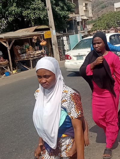 Two women stand on a paved road, with a car visible behind them. The woman in the foreground wears a white headscarf and patterned dress, while the other wears a black headscarf and bright pink outfit. A small stall or shop lines the road, overflowing with assorted items. It is located nearby a lush green tree.
