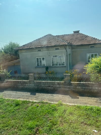 The image depicts a single-story house with a tiled roof, situated in Kardam, Bulgaria. The house is painted a light gray color and features several windows, some of which are covered with blinds. A chimney is visible on the roof. In front of the house, there is a low stone wall topped with an ornamental metal fence. Yellow flowers are blooming in bushes near the fence and the house. To the left of the house, a smaller structure with a blue door is partially visible. The foreground of the image shows a grassy area next to a paved path. The sky is clear and blue, indicating a sunny day.