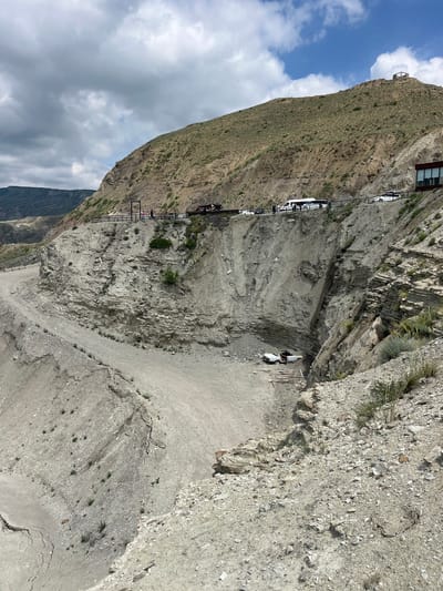 The image depicts a steep, rocky cliffside in Chirkey, Russia. A narrow road is carved into the cliff, with several cars parked along it. A metal walkway with railings runs parallel to the road, seemingly providing access to a building or viewing point at the top of the cliff. Two figures are visible near a vehicle at the edge of the cliffside, suggesting human activity. The environment is arid and mountainous, with limited vegetation. The sky is partially cloudy.