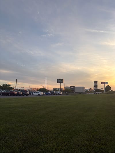 This is a wide-angle, horizontal, full-shot, eye-level photograph of a commercial area at dusk, likely in the city of Halfway, United States of America. The sky is a mix of pale blue and soft orange, with scattered wispy clouds. The sun is low on the horizon, casting a warm glow over the scene. In the foreground, a lush green lawn stretches across the bottom of the frame. Behind the lawn, a parking lot is filled with various cars, including sedans and SUVs. Several streetlights are visible throughout the parking lot. In the background, commercial buildings are lined up, featuring prominent signage for businesses such as Olive Garden, Noodles & Company, and Starbucks. A tall, multi-tenant sign also stands out. The overall atmosphere is calm and serene, characteristic of the end of a day. There are no people or animals clearly visible in the photograph.