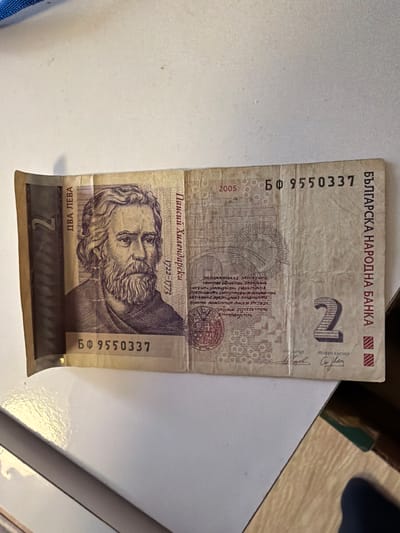 The image shows a two Bulgarian Leva banknote. The banknote features Paisiy Hilendarski, a Bulgarian clergyman and writer and an important figure in the Bulgarian National Revival, as well as the text "БЪЛГАРСКА НАРОДНА БАHKА" (Bulgarian National Bank) at the top. The text "ДВА ЛЕВА" (Two Leva) is at the bottom, and the years "1722-1773" are printed above Hilendarski's image. The serial number "БФ 9550337" is printed vertically on both sides. The banknote is placed on a white surface. The image provides no information about a location in the city of Radomir, Bulgaria.