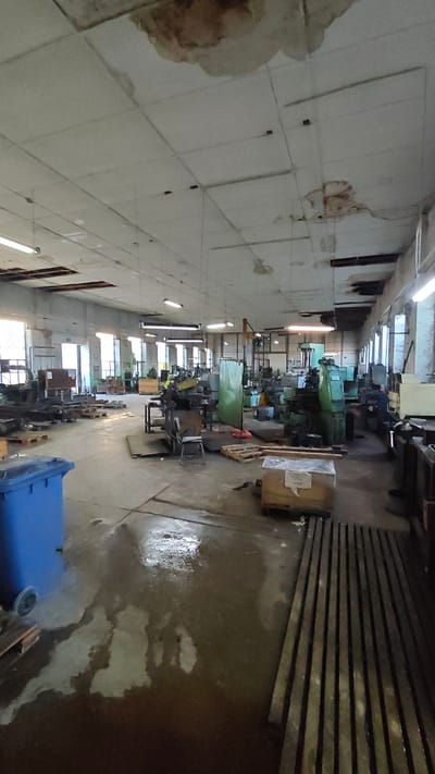 The image depicts the interior of an industrial workshop or factory located in Goczałków, Poland. The space is large, with a concrete floor that appears wet and reflects light in several areas. The ceiling is white, with visible water damage and staining in multiple places. Fluorescent lighting fixtures are installed on the ceiling, some of which are illuminated.

The workshop is filled with various heavy machinery, likely metalworking equipment, such as lathes and milling machines, predominantly colored in green and gray. Many of these machines are positioned throughout the room. Wooden pallets are scattered on the floor, some with materials or boxes placed on them. A blue industrial waste bin is visible on the left side of the frame. In the background, large windows allow natural light to enter the space. The overall impression is one of a functional but somewhat neglected industrial environment. There are no people visible in the image.