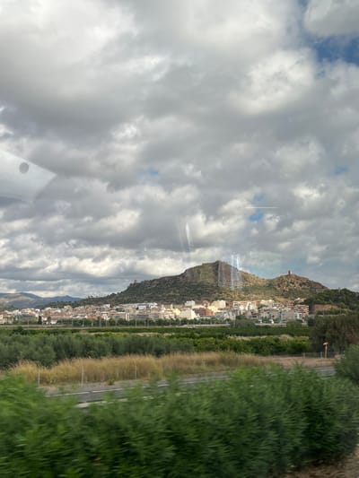 The image depicts a panoramic landscape of Almenara, Spain, observed under a cloudy sky with intermittent blue patches. The upper half of the frame is dominated by a dense formation of grey and white clouds, with a visible circular reflection in the upper left quadrant and several vertical light streaks across the frame, consistent with reflections off a window.

In the background, a series of hills and mountains rise, with the central and most prominent hill exhibiting rugged, exposed rock formations and sparse green vegetation. Situated at the base and extending partway up these hills is the city of Almenara, characterized by numerous light-colored buildings with varied rooflines. Two distinct, brown-toned tower structures, likely historical fortifications (e.g., Almenara Castle remnants), are visible on separate peaks/ridges of the main hill, one to the left of the central peak and another to the right.

The midground features cultivated land, consisting of rows of dense green trees, indicative of orchards or groves, interspersed with patches of dry, lighter-colored vegetation. A two-lane road or highway, demarcated by white lines and a thin barrier or guardrail, runs horizontally through this area.

The foreground is occupied by blurred green foliage, likely shrubs or bushes, suggesting proximity to the camera and motion blur, consistent with a photograph taken from a moving vehicle. No individuals or specific human activities are discernable within the scene.