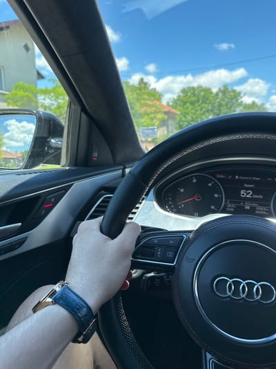 The image shows a first-person perspective of someone driving an Audi. The car's dashboard is visible, with a speedometer displaying 52 km/h. The driver's hand is on the steering wheel, and a watch with a blue strap is on their wrist. The exterior view through the windshield shows a sunny day with blue skies and clouds, and trees and buildings. Based on the available information and the visual cues in the image, the location is likely the city of Marten, Bulgaria.