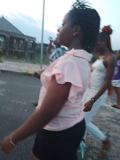 A medium shot, taken outdoors, features a group of people walking on a paved road. The main subject is a woman in the foreground, walking in profile. She wears a light pink collared shirt with short, ruffled sleeves, paired with black shorts. A gold bracelet is visible on her wrist. Her hair is braided, and a possible hair accessory adorns her head.

In the background, another woman walks in a white outfit. Further back, additional figures are visible, including a person holding a beverage. An unfinished building is visible in the blurred background, along with a utility pole. The sky is overcast, and the overall lighting suggests either dusk or dawn. The scene is located in Umueshi, Nigeria.