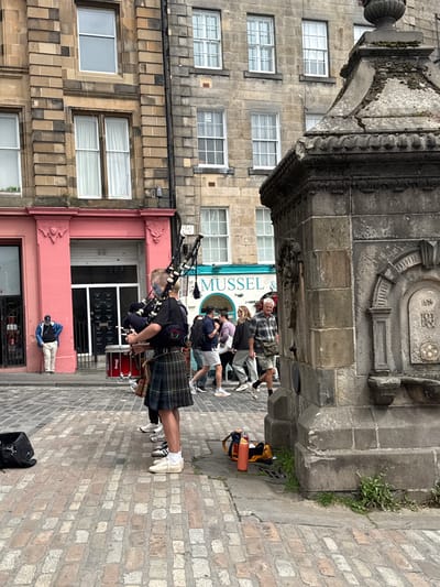 The image shows a street scene in the City of Edinburgh, United Kingdom. A bagpiper dressed in traditional Scottish attire, including a kilt, is playing bagpipes in the foreground. A red drum is positioned next to the bagpiper. Several people are walking in the background. Buildings with stone facades and windows line the street. A stone fountain is visible on the right side of the image. The ground is paved with cobblestones.