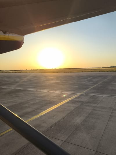 The image shows an airplane wing and fuselage casting a shadow over an airport tarmac under a bright, setting sun. The tarmac is made of concrete slabs and marked with yellow lines. In the distance, there are fields and trees. The sky is clear and transitions from a pale blue to a warm yellow near the horizon. The scene is likely at Schönefeld Airport (now Berlin Brandenburg Airport) in Schönefeld, Germany, given the context of an airport setting with an airplane. There are no people or immediate actions depicted.