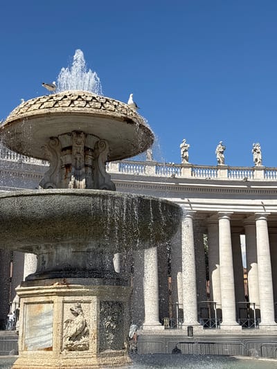 The image captures a detailed view of the Fountain of Neptune in St. Peter's Square, Città del Vaticano, Vatican City. A tiered stone fountain is prominently featured in the foreground, with water actively cascading down its levels, creating a dynamic play of light and shadow. Two seagulls are perched atop the upper tier of the fountain, silhouetted against a clear, bright blue sky.
Behind the fountain, the colonnade of St. Peter's Square stretches across the background. The Bernini-designed colonnade is characterized by its towering marble columns, supporting a balustrade topped with numerous saint statues. Three of these statues are clearly visible on the upper balustrade, depicted in classical drapery. The overall setting is one of grandeur and historical significance, with the vibrant blue sky and flowing water adding a sense of life to the ancient architecture.