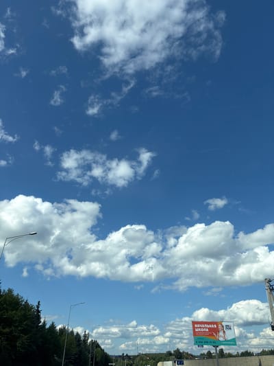 The image, taken in Obushkovo, Russia, captures a daytime scene with a vast expanse of blue sky dotted with fluffy white clouds. In the foreground, a line of dark green trees borders a road, with several streetlights visible. To the right, a billboard displays an advertisement for "НАЧАЛЬНАЯ ШКОЛА" (Primary School) featuring a young girl and contact information. The overall environment suggests a suburban or rural setting under a clear, sunny sky.