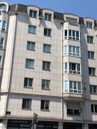 This is a medium-shot, tilted, exterior photograph of a modern building with a light-colored, tiled facade. The building appears to be multi-story, with numerous windows of various sizes and styles. Some windows are large and panoramic, while others are smaller and rectangular. Several windows are open, suggesting the building is inhabited or in use. The architectural style is contemporary, characterized by clean lines and geometric shapes.

The building is situated outdoors, under a clear, bright blue sky, indicating daytime with fair weather. The sunlight casts distinct shadows on the facade, highlighting the architectural features and the grid pattern of the tiles. The angle of the shot suggests the photographer is looking up at the building from a low vantage point or that the building itself is leaning or angled.

Visible on the side of the building is a traffic light, a green pedestrian crossing signal, and a pole with vertical text. The text on the pole reads "01 42 42 02 02." There is also a small rectangular plaque on the wall below the traffic signal.

The scene is devoid of people, focusing entirely on the architecture and its surroundings. The overall impression is one of urban development and modern design in Courbevoie, France.
