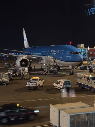 At night, a large KLM Asia airplane is parked at Aeropuerto Internacional Ezeiza, Argentina. The aircraft is blue and white, with the KLM logo and "KLM ASIA" clearly visible on its fuselage and tail. Several service vehicles are positioned around the plane, including a white pickup truck with flashing amber lights, a white van, and a large security vehicle labeled "BRINKS." Ground crew members, visible as small figures, are attending to the aircraft and vehicles. In the foreground, a black car is blurred due to motion, suggesting it is moving across the tarmac. The scene is illuminated by the aircraft's lights, the service vehicles' lights, and the ambient lighting of the airport. A jet bridge connects to the airplane. The overall impression is one of routine airport operations occurring at night.