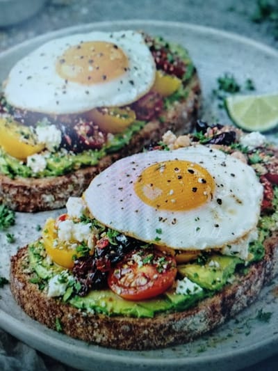 The image shows two pieces of avocado toast topped with fried eggs, tomatoes, feta cheese, and herbs. The toast is on a grey plate. A lime wedge is visible to the right of the plate. The image does not contain any information about the city of Yashkino, Russia.