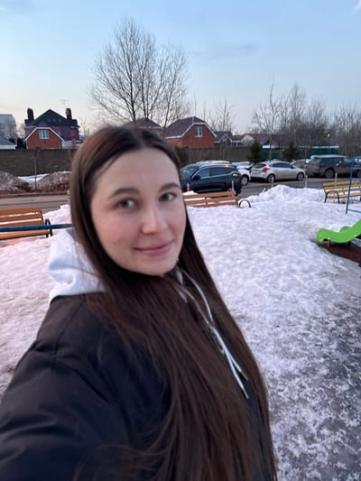 The scene is an outdoor environment in Akulovo (Gp Kubinka), Russia, characterized by patchy snow and ice cover on the ground. A female subject occupies the foreground, facing the camera. She presents with long, dark brown hair and is dressed in a dark jacket over a white hooded sweatshirt. Her facial expression is a slight smile, with her eyes directed slightly left of the camera's axis. To the subject's left, a portion of a wooden slatted bench with blue metal supports is visible. To her right, part of a green plastic playground slide is observed. In the midground, a paved area serves as a parking space for several vehicles, including a dark SUV and a light-colored sedan. The background features a low earth-toned retaining wall, beyond which are multiple detached residential structures with pitched roofs and numerous bare deciduous trees. The sky is pale blue, suggesting ambient light conditions consistent with dawn or dusk.