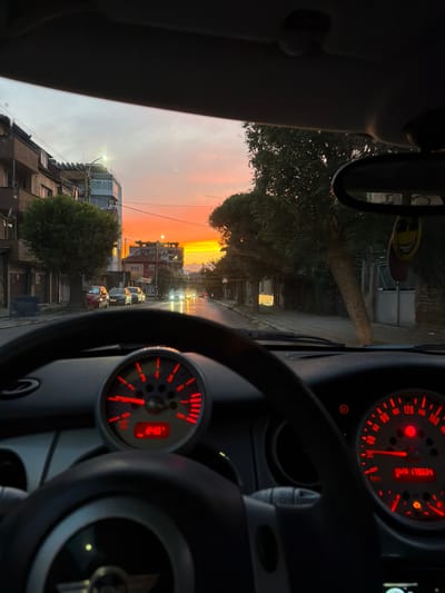 The image is taken from the interior of a car, looking out through the windshield at a city street scene during sunset. The sky is ablaze with orange and yellow hues. Buildings line both sides of the street, with trees interspersed. Several cars are visible on the road, and streetlights are illuminated. The car's dashboard is also visible, featuring a steering wheel, a speedometer, and a tachometer with red needles and illuminated numbers. A smiley face air freshener hangs from the rearview mirror. The scene suggests the car is either parked or moving slowly through the city. The location is specified as Dupnitsa, Bulgaria.