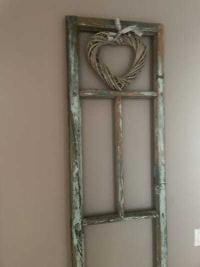 This is a horizontal, medium-shot, eye-level photograph of a decorative ladder leaning against a plain, taupe-colored wall. The ladder is made of aged, weathered wood, with a distressed finish that shows patches of teal paint and exposed wood grain. It is constructed with two vertical supports and three horizontal rungs, forming three rectangular sections.

Hanging on the leftmost section of the ladder is a heart-shaped wreath made of woven natural material, possibly twigs or straw. The wreath is tied to the ladder with a simple white ribbon or string. The lighting suggests an indoor setting with soft, diffused light, and there are no indicators of time of day or weather. The overall mood is rustic and decorative. There is no visible text in the image. The location context provided is Мухавецкі сельскі Савет, Belarus.