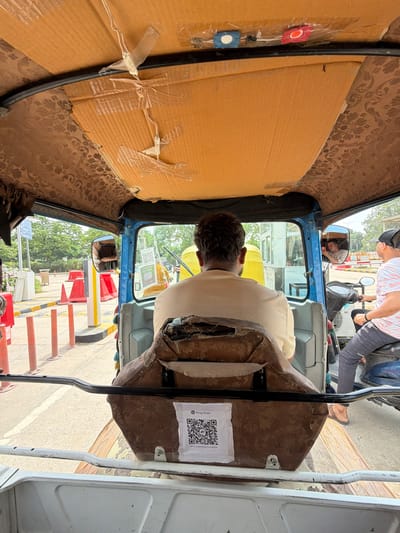 The image captures the interior of a blue auto-rickshaw in Aurangabad, India, looking forward. The auto-rickshaw's interior ceiling is covered with orange cardboard and brown patterned fabric. A QR code for "King Khan" is visible on the backrest of the seat. In the front, a driver wearing a cream-colored shirt is visible from the back, and the passenger side mirror reflects a person holding a phone. Outside the auto-rickshaw, traffic barriers and orange cones line a road, suggesting a toll booth or checkpoint. Another auto-rickshaw, this one yellow, is visible in the distance. To the right, a person on a scooter wearing a white and pink striped shirt and a black cap is also in view. The environment appears to be an outdoor setting with trees and buildings in the background.