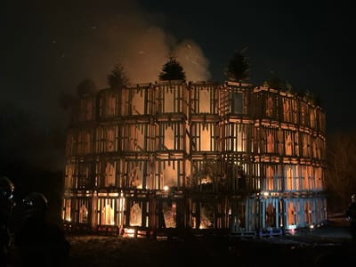 The image captures a large, multi-tiered bonfire structure burning brightly against a dark night sky. The primary subject is a massive, cylindrical or circular tower-like construction made almost entirely of wooden pallets. This structure is several stories high, appearing to have at least three to four distinct tiers, each formed by stacking pallets to create an intricate, grid-like pattern of open compartments.

Inside these wooden compartments, and particularly at the base and various levels of the structure, a substantial fire is actively burning. The flames emit a vivid orange and yellow glow, illuminating the wooden framework from within and casting warm light onto the surrounding area. Smoke billows upwards from the left side of the structure, partially obscuring the sky, and numerous small sparks and embers can be seen rising into the darkness.

On the very top tier of the wooden structure, several small evergreen trees, resembling Christmas trees, are positioned. Some of these trees appear to be illuminated by the flames below or are themselves beginning to burn, contributing to the overall inferno.

In the lower-left foreground, two silhouetted figures are visible, observing the burning structure. Their forms are dark against the bright fire, but they appear to be wearing helmets, suggesting they might be event organizers, safety personnel, or firefighters.

The scene takes place outdoors at night, with no other discernible light sources apart from the fire itself. The background is dark, with faint outlines of bare trees or brush just beyond the structure on the left. The overall impression is one of a large-scale, controlled bonfire event, creating a dramatic and visually striking spectacle of light, heat, and smoke. No text is visible in the image.