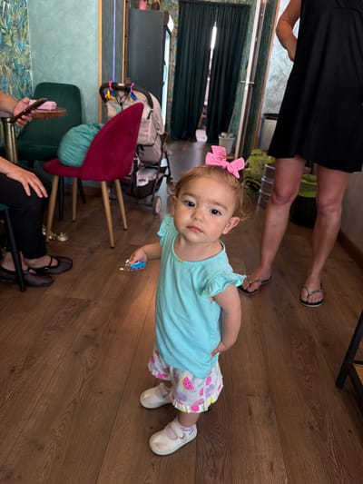 Captured from a high-angle perspective, the image reveals the interior of what seems to be a mobile home or trailer. The flooring is a dark-stained hardwood, adding a warm tone to the scene. In the foreground, a toddler, adorned with a pink hair bow, gazes directly at the camera. She wears a light blue shirt and patterned shorts, with white shoes completing her attire.

An adult stands partially visible at the top of the frame, sporting a black dress and sandals. Another individual is seated in the lower left, wearing black trousers and Mary Jane shoes, with hands visible holding a phone or similar device.

The backdrop of the scene shows signs of a compact living space, including an array of items like a backpack, blankets, and what appears to be a child’s stroller. The overall setting suggests a lived-in, somewhat cramped environment.

While the image’s metadata isn't available, the request includes the city of Salou, Spain. Thus, it's assumed this scene takes place inside a trailer or mobile home located in Salou, Spain, providing a sense of context to the location.