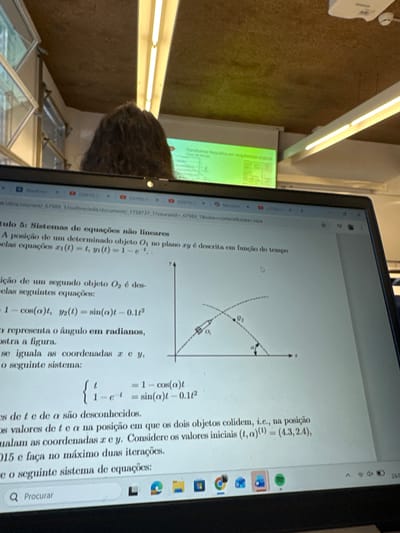 The scene is set indoors within an academic building, likely a classroom or lecture hall at the University of Minho in Guimarães, Portugal.
In the foreground, a black laptop displays a document in Portuguese. The title "Capítulo 5: Sistemas de equações não lineares" (Chapter 5: Systems of nonlinear equations) is visible. The document details a mathematical problem involving the trajectories of two objects, O1 and O2, described by time-dependent equations ($x_1(t)=t, y_1(t)=1-e^{-t}$ and $x_2(t)=1-\cos(\alpha)t, y_2(t)=\sin(\alpha)t-0.1t^2$). A corresponding Cartesian graph illustrates these trajectories, showing an angle 'α' and two objects at various points. The text specifies finding unknown values of 't' and 'α' for the objects' collision, providing initial values $(t, \alpha)^{(1)} = (4.3, 2.4)$ and limiting iterations to two. The laptop's taskbar is visible at the bottom, displaying icons for Windows, Google Chrome, Microsoft Edge, Microsoft Word, and Spotify, along with a "24/0" date stamp.
In the mid-ground, the back of a person's head with dark, curly hair is visible, positioned directly in front of the laptop. This individual faces away from the camera.
The background features a white projector screen displaying a presentation slide with green and white content, including text and possibly diagrams. A white projector unit is mounted on the textured, likely concrete, ceiling above the screen. Linear fluorescent light fixtures illuminate the room from the ceiling. A window or glass partition is partially visible on the far left.