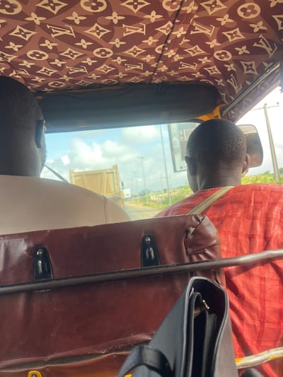 The image is taken from inside an auto-rickshaw, likely in Karum, Nigeria. Two individuals are visible. One person is in the foreground, with their back to the camera, wearing a red, textured garment. They have a beaded necklace. Another person is visible through the windshield, with their head turned away from the camera, wearing a dark, textured outfit. The interior of the auto-rickshaw is decorated with a repeating pattern of what appears to be the Louis Vuitton monogram. A black bag is placed on a brown leather seat. Outside the vehicle, a cloudy sky is visible, suggesting it is daytime. A yellow dump truck is seen in the distance. The overall impression is of a journey or commute.