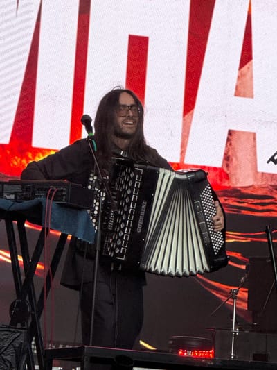 A man with dark, shoulder-length hair and glasses is playing an accordion on a stage. He is wearing a dark jacket and dark pants. He is smiling and looking to his right. The accordion is black and white with silver buttons. To his left, there is a keyboard on a stand with red wires. In the background, there is a large screen with red and white diagonal stripes. The stage appears to be outdoors, as indicated by the lighting and the general atmosphere. There is no discernible information in the image to suggest a specific location within Lagunovo, Russia.