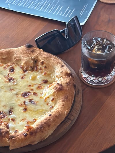 On a wooden table, a pizza sits on a round wooden serving board. To the left are black sunglasses and a menu. A glass of dark liquid with ice rests on a coaster above the pizza. This scene is likely set in Tegal Suci, Indonesia.