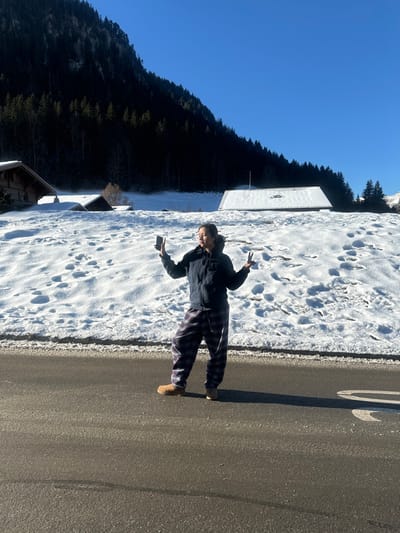 A full-body image depicts an individual standing on an asphalt road in Saanen, Switzerland, against a snow-covered alpine landscape under a clear blue sky. The individual, appearing to be female, is positioned centrally, wearing a dark hooded jacket, plaid pajama-style pants in shades of purple and blue, and light brown boots. She holds a black smartphone vertically in her raised left hand, directed towards herself, consistent with taking a selfie. Her right hand is raised, forming a "V" or peace sign. Her head is tilted slightly right, with her eyes partially closed. Behind the individual, a snow-covered hillside extends upwards, displaying numerous footprints. On this slope, two traditional wooden chalets with snow-covered roofs are visible on the left side, with another smaller structure further to the right. In the far background, a steep mountain face is covered with dense, dark green coniferous trees, partially shadowed. The road in the foreground is dark asphalt, with a faint white marking visible to the right of the individual's feet. Bright sunlight illuminates the scene, casting a distinct shadow of the individual on the road to her right.
