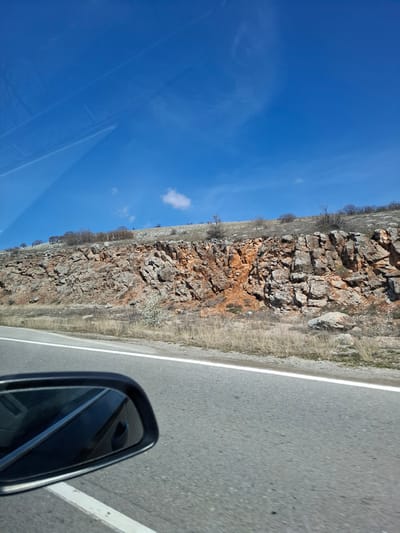 The image depicts a view from a moving vehicle on a road near Bogyovtsi, Bulgaria. The road is asphalt and has a white line marking the edge. To the left of the road is a rocky, barren hillside with sparse vegetation. The sky is clear and blue, with a few small clouds. The side mirror of the car is visible in the lower right corner of the frame.