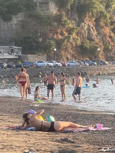 The image captures a detailed scene on a beach in Sestri Levante, Italy, characterized by a coastal environment with a bay, a paved road, and a steep, vegetated hillside.
In the foreground, a female subject is lying prone on a dark mat or towel on the sandy-pebbly beach. She wears a dark two-piece swimsuit and displays tattoos on her right arm and lower back. Her dark hair is pulled back. Nearby, a pair of purple sandals, a yellow item, and a green bag are on the sand. To her right, a pink bucket and other colorful beach toys are placed on a light-colored towel.
The midground features numerous individuals in and around the calm, shallow bay water. At the water's edge, a woman in a red bikini is walking away from the camera, while another person is partially visible next to her. A man with a beard and dark swim shorts stands with a woman in a dark bikini. Another man in dark swim shorts with white stripes is positioned near them. A woman in a dark bikini sits on the sand at the water's edge. Multiple other individuals, including adults and children, are wading, swimming, or using inflatable rings further out in the water. Small waves ripple on the water's surface.
Behind the beachgoers, a low, grey stone wall separates the beach from a paved area serving as a road or parking lot. Approximately ten to twelve passenger vehicles of various models and colors (including silver, blue, red, and black) are parked in a row along this wall. Two tall poles, likely streetlights with spherical white lamps, are visible along the roadside.
The background is dominated by a steep, rugged hillside or cliff covered with sparse, dry-looking vegetation and some greener bushes. Stone structures, including what appear to be retaining walls and parts of buildings, are discernible on the hillside. A multi-story building with visible windows is situated at the base of the hill near the parking area. The lighting suggests late afternoon or early evening.