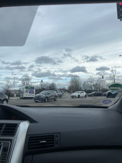 The image presents a forensic view from inside a vehicle at a multi-lane road intersection in Illkirch-Graffenstaden, France, at the crossing of Route de Lyon (D468) and Rue de l'Industrie / Avenue de Strasbourg. The weather is overcast with a mix of grey and blue sky.
The lower foreground is occupied by the dark grey dashboard of the observer's vehicle, featuring air vents and control panels. The upper part of the windshield shows a partial view of a red-illuminated traffic light on the right and several circular stickers adhered to the glass.
At the intersection, a dark grey Renault Captur SUV, bearing French license plate DL-463-MY, is centrally positioned. To its right, a white Volkswagen Scirocco is visible, followed by a black Mercedes-Benz C-Class sedan and another dark sedan further right. A black SUV is partially visible on the far left. A white public transport bus, marked with "C7 Sud" and "T'RIGA" branding, is situated in the left lane behind the Renault Captur. A green canopy, indicative of a gas station, is visible behind the bus. Bare trees, traffic lights, and streetlights line the intersection and surrounding areas. A distinctive tall, modern building, likely part of the Parc d'Innovation, is visible in the distant background. Vehicles are either stopped at the red light or preparing to move through the intersection.