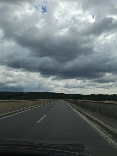 The image shows a view from a car driving on a highway bridge, likely in or near Podolí I, Czechia, on a cloudy day. The road is asphalt with a white line marking the lane. On either side of the road are concrete barriers topped with decorative spherical elements. Beyond the barriers, a dense green forest lines the horizon. The sky is filled with dramatic, dark gray clouds with patches of lighter gray and a small glimpse of blue sky. The overall impression is one of a slightly overcast and moody landscape.