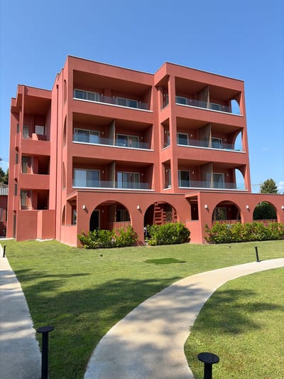 A modern, salmon-colored building with multiple balconies and arched ground-floor entrances stands under a clear blue sky. The building has a geometric design with recessed balconies and glass railings. In front of the building, a well-maintained green lawn is bordered by a curved concrete pathway. Several black, low-profile lampposts are spaced along the pathway. Lush green bushes are planted in front of the arched entrances on the ground level. The scene suggests a resort or hotel in a sunny, possibly coastal, location. This building is located in Pallini, Greece.