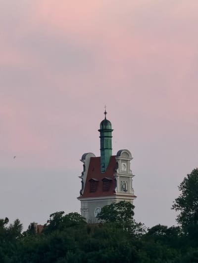 The image shows a building in Sopot, Poland, with a distinctive turret topped by a green dome and a cross. The building has a red-tiled roof and ornate architectural details. Trees partially obscure the lower part of the building, and a soft, pinkish sky fills the background. A small bird is visible in the sky to the left of the turret.