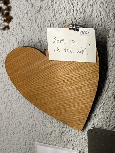 A wooden heart-shaped object hangs on a textured, light-colored wall. A small white card is attached to the top of the heart with a black binder clip. Handwritten on the card in black ink are the words "love is in the air!" Below these words, in the upper right corner of the card, is a handwritten price tag that reads "19.95". A blurred, dark object is visible in the lower right corner of the image. The scene is indoors, with no indication of the specific location within St. Märgen, Germany.