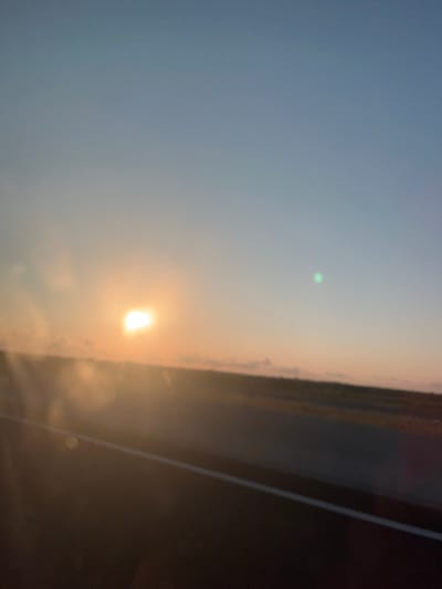 The image captures a blurred view of a sunset or sunrise over a flat, grassy landscape. The sky transitions from a pale blue at the top to hues of orange and pink near the horizon. The sun is a bright, glowing orb, partially obscured by hazy clouds, located in the upper left quadrant of the image. A faint green light, possibly a lens flare or atmospheric phenomenon, is visible in the upper right portion of the sky. In the foreground, a dark, indistinct band suggests a road or a paved surface, marked by a faint white line. The overall scene is hazy and appears to be taken from a moving vehicle. The location is identified as Lesi Community, Georgia.